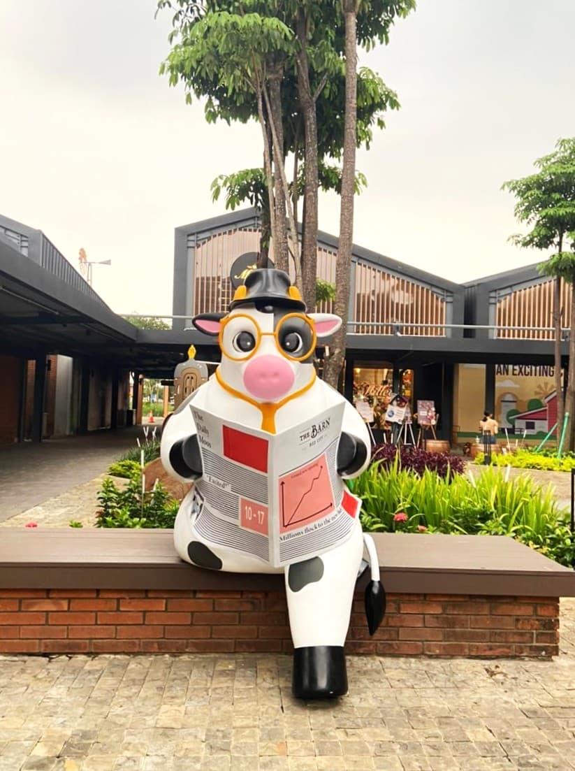 Patung sapi raksasa berkacamata dan bertopi sedang membaca koran di bangku di The Barn, BSD City, Indonesia